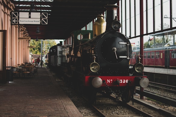 A vintage steam locomotive is positioned on train tracks inside a station with large glass windows. The locomotive has a distinct circular front and metallic components, with a visible number plate reading 'No 326' on the front. To the left, there is an area with outdoor seating and a sign labeled 'KAARTVERKOOP/KASSA,' indicating a ticket booth. There are also signs of another train visible through the windows on the right side.