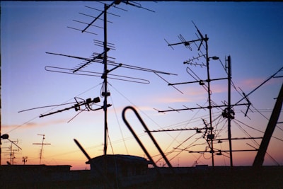 A variety of television antennas are silhouetted against a colorful sunset sky, with hues of orange, purple, and blue. The antennas are mounted on rooftops and connected by several cables, creating an intricate network of lines against the fading light.