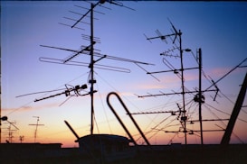 A variety of television antennas are silhouetted against a colorful sunset sky, with hues of orange, purple, and blue. The antennas are mounted on rooftops and connected by several cables, creating an intricate network of lines against the fading light.