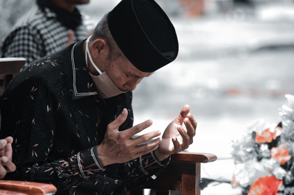 An elderly person is sitting with a contemplative expression, hands raised in a prayer gesture. The person is wearing a black cap and a face mask. The clothing has intricate patterns, and there is a blurred background with muted colors, alongside some vibrant flowers in the foreground.