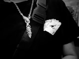 A close-up of a person wearing a black t-shirt with a graphic of a hand holding playing cards, featuring the text 'Off-White 2020'. A metallic chain with a decorative pendant hangs around the neck, and a black strap with a buckle is visible.