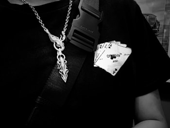 A close-up of a person wearing a black t-shirt with a graphic of a hand holding playing cards, featuring the text 'Off-White 2020'. A metallic chain with a decorative pendant hangs around the neck, and a black strap with a buckle is visible.