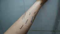 A forearm with droplets of water and words written in different orientations in black ink. Words include 'sadness', 'loneliness', and 'melancholia'. The background appears to be a tiled wall.