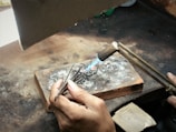 Two hands are working on a piece of jewelry or metal with a small blowtorch. The workspace includes a brick for heat resistance and various metal fragments. The person is holding a pair of tweezers to manipulate the object under the flame.