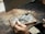 Two hands are working on a piece of jewelry or metal with a small blowtorch. The workspace includes a brick for heat resistance and various metal fragments. The person is holding a pair of tweezers to manipulate the object under the flame.