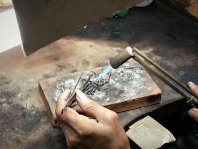Two hands are working on a piece of jewelry or metal with a small blowtorch. The workspace includes a brick for heat resistance and various metal fragments. The person is holding a pair of tweezers to manipulate the object under the flame.