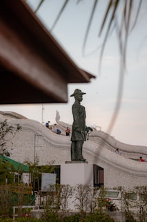 A large bronze statue of a historical figure wearing a traditional hat stands prominently on a white pedestal. In the background, a group of people is seen on a decorative structure with white, curved architecture, possibly a temple or cultural building. The setting includes greenery in the foreground with a partly cloudy sky.
