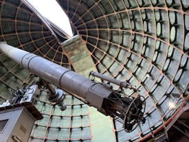 A large telescope is positioned inside a dome with a geometric, honeycomb-like ceiling structure. The telescope is metallic, with varying sections and components, standing on a base that appears mechanical and complex. Light enters from an opening at the top, highlighting the intricate design and construction of the telescope and the surrounding dome.