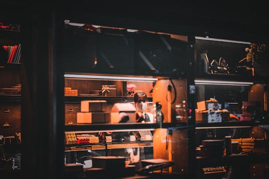 A warmly lit shop interior with wooden shelves filled with various boxes and a person wearing a mask, focused on a task behind a glass window. The glow from the lights creates a cozy atmosphere, with reflections adding depth to the scene.