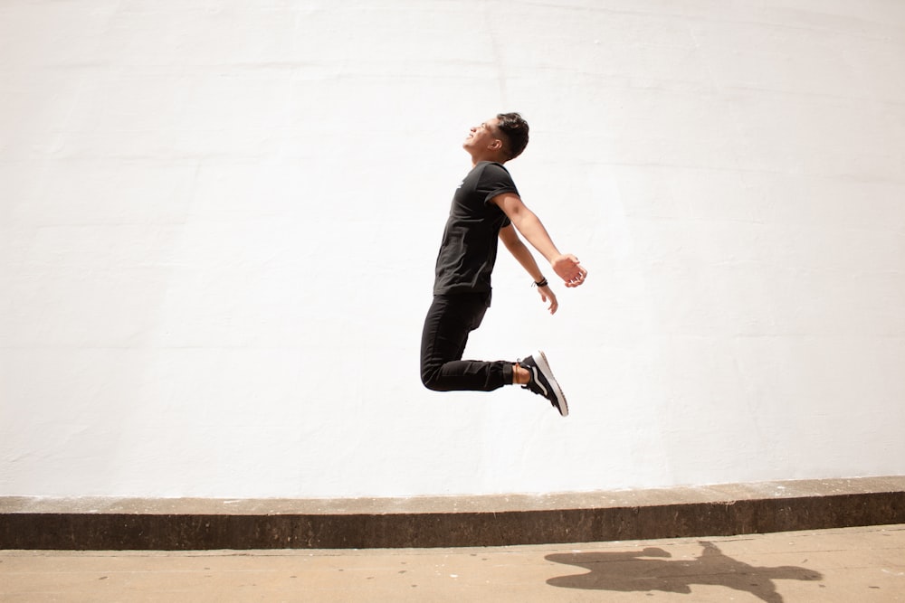 homme en veste noire et pantalon noir sautant sur un mur de béton blanc pendant la journée