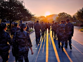 A group of people in military uniforms walk along a paved path during sunset. The sun is low on the horizon, casting long shadows and creating a warm glow across the scene. Trees line the path, and a modern building is visible in the background.