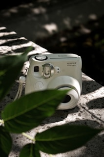 A white Fujifilm instant camera rests on a textured stone surface, partially obscured by vibrant green leaves. Dappled sunlight creates patterns of light and shadow, adding depth to the scene.