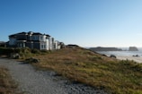 A large coastal house is situated near a path lined with grass and rocks, overlooking a sandy beach with distant rock formations and a clear blue sky.