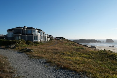 A large coastal house is situated near a path lined with grass and rocks, overlooking a sandy beach with distant rock formations and a clear blue sky.