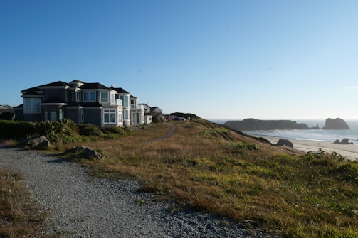 A large coastal house is situated near a path lined with grass and rocks, overlooking a sandy beach with distant rock formations and a clear blue sky.
