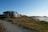 A large coastal house is situated near a path lined with grass and rocks, overlooking a sandy beach with distant rock formations and a clear blue sky.