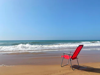 A vibrant red beach chair faces the ocean on a sandy beach, with gentle waves rolling in under a clear blue sky.