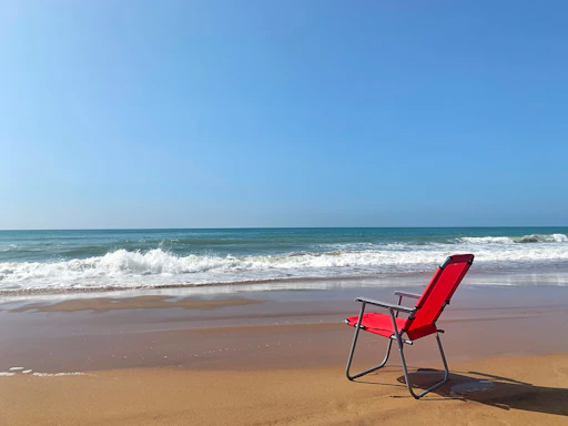 A vibrant red beach chair faces the ocean on a sandy beach, with gentle waves rolling in under a clear blue sky.