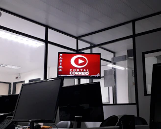The image depicts an office space with several computer monitors on desks. A red digital display on the wall reads 'Portal Correio'. The room has glass partitions and a modern, streamlined design. Overhead fluorescent lights provide illumination.