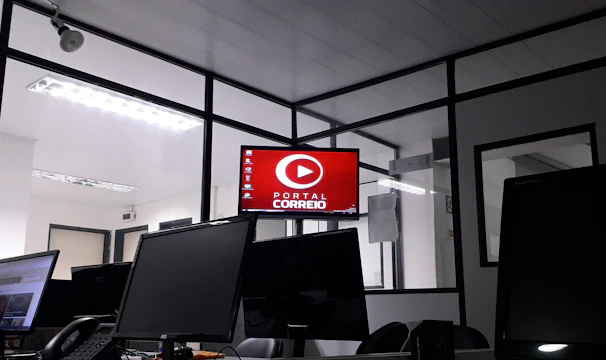 The image depicts an office space with several computer monitors on desks. A red digital display on the wall reads 'Portal Correio'. The room has glass partitions and a modern, streamlined design. Overhead fluorescent lights provide illumination.