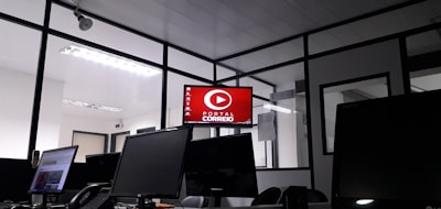 The image depicts an office space with several computer monitors on desks. A red digital display on the wall reads 'Portal Correio'. The room has glass partitions and a modern, streamlined design. Overhead fluorescent lights provide illumination.