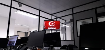 The image depicts an office space with several computer monitors on desks. A red digital display on the wall reads 'Portal Correio'. The room has glass partitions and a modern, streamlined design. Overhead fluorescent lights provide illumination.
