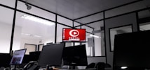 The image depicts an office space with several computer monitors on desks. A red digital display on the wall reads 'Portal Correio'. The room has glass partitions and a modern, streamlined design. Overhead fluorescent lights provide illumination.