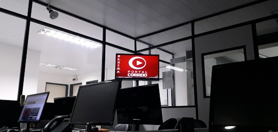 The image depicts an office space with several computer monitors on desks. A red digital display on the wall reads 'Portal Correio'. The room has glass partitions and a modern, streamlined design. Overhead fluorescent lights provide illumination.
