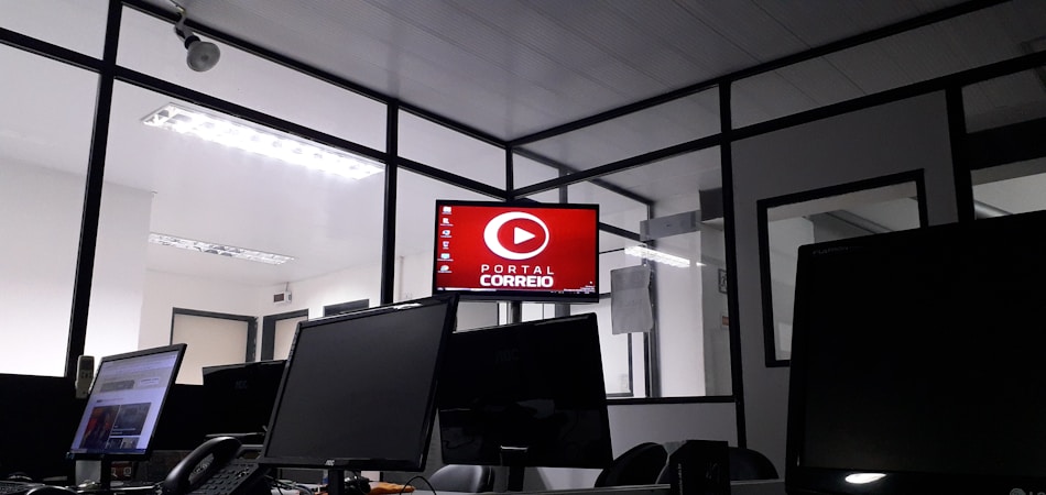 The image depicts an office space with several computer monitors on desks. A red digital display on the wall reads 'Portal Correio'. The room has glass partitions and a modern, streamlined design. Overhead fluorescent lights provide illumination.