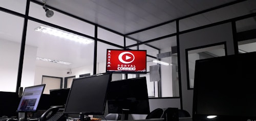 The image depicts an office space with several computer monitors on desks. A red digital display on the wall reads 'Portal Correio'. The room has glass partitions and a modern, streamlined design. Overhead fluorescent lights provide illumination.