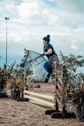 A person is riding a white horse as it jumps over a wooden hurdle covered with dried leaves in an outdoor equestrian setting. The rider is dressed in a helmet and casual riding gear.