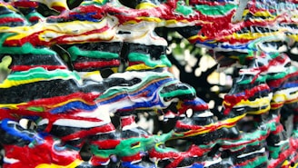 An abstract sculpture featuring thick, intertwined layers of brightly colored paint in red, green, yellow, white, blue, and black, creating a visually chaotic and textured pattern.