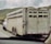 A livestock trailer is being towed by a white pickup truck on a highway. The trailer, constructed of metal, has multiple ventilation slats on its sides. It is equipped with several wheels and appears to be designed for transporting animals. The surrounding landscape includes green hills under a cloudy sky.