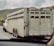A livestock trailer is being towed by a white pickup truck on a highway. The trailer, constructed of metal, has multiple ventilation slats on its sides. It is equipped with several wheels and appears to be designed for transporting animals. The surrounding landscape includes green hills under a cloudy sky.