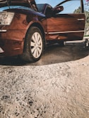A car with its front hood and driver-side door open, parked on a rough, textured pavement. The vehicle appears to be in a stationary position, with a clear view of the front left tire and alloy wheel. Sunlight casts shadows on the pavement, highlighting the car’s glossy maroon paint. In the background, there are trees and power lines visible under a blue sky.