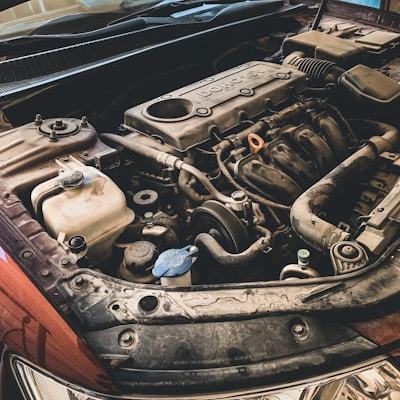 A detailed view of a car engine, showing various components such as the valve cover with the word 'DOHC', pipes, hoses, and the belt system. The surrounding parts have some dirt and dust, indicating possible wear or everyday use.