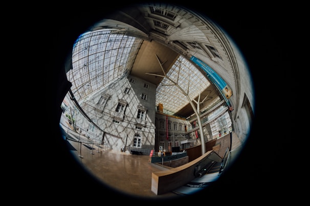 An architectural interior view captured through a fisheye lens, creating a circular, distorted effect. The scene includes tall, ornate building facades with many windows, large skylights with a grid structure, and a grand lobby area. The lighting casts intricate shadows across the surfaces, and there are modern elements like an escalator and informational banners.