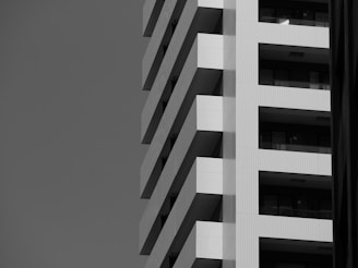 A black and white photograph of a modern building showcasing its geometric architecture. The building features a series of balconies, each with a sharp, angular design.