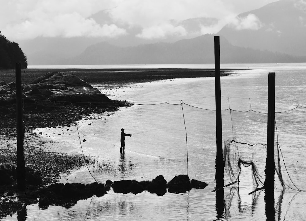 grayscale photo of person standing on rock near body of water