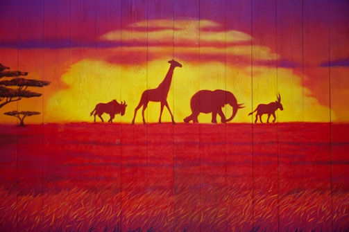 Silhouettes of animals including a giraffe, elephant, and wildebeest appear against a vibrant sunset, with warm orange, red, and yellow tones dominating the scene. The landscape has an African savanna theme, with acacia trees visible on the left.