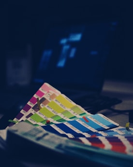Several color swatches are spread out in the foreground with their vibrant hues visible. Behind them, there is a blurred laptop screen displaying an unclear interface with hazy blue light illuminating around. The overall scene appears to be dimly lit.
