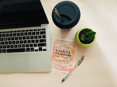 A workspace setup featuring a laptop with a visible keyboard on the left, a black drinking cup with a straw at the top right, a small potted plant in a green pot, a motivational card with calligraphy emphasizing vision and creation, and a transparent pen lies next to it.