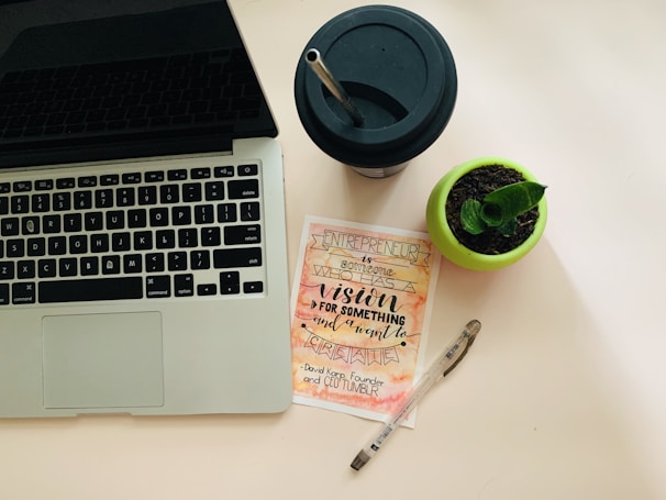 A workspace setup featuring a laptop with a visible keyboard on the left, a black drinking cup with a straw at the top right, a small potted plant in a green pot, a motivational card with calligraphy emphasizing vision and creation, and a transparent pen lies next to it.