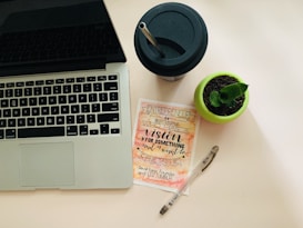 A workspace setup featuring a laptop with a visible keyboard on the left, a black drinking cup with a straw at the top right, a small potted plant in a green pot, a motivational card with calligraphy emphasizing vision and creation, and a transparent pen lies next to it.