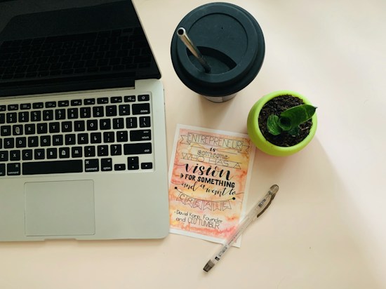 A workspace setup featuring a laptop with a visible keyboard on the left, a black drinking cup with a straw at the top right, a small potted plant in a green pot, a motivational card with calligraphy emphasizing vision and creation, and a transparent pen lies next to it.
