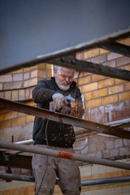 A person is using a grinder to cut through metal beams, producing bright sparks. They are working against a backdrop of a brick wall, wearing protective eyewear and gloves.