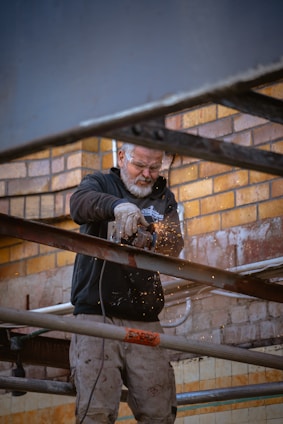 A person is using a grinder to cut through metal beams, producing bright sparks. They are working against a backdrop of a brick wall, wearing protective eyewear and gloves.