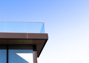 A corner of a modern building featuring a glass railing on an upper balcony against a clear blue sky. The architecture showcases sleek lines with a minimalist design.