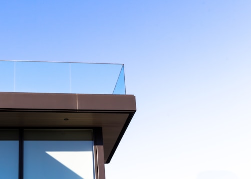 A corner of a modern building featuring a glass railing on an upper balcony against a clear blue sky. The architecture showcases sleek lines with a minimalist design.
