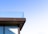 A corner of a modern building featuring a glass railing on an upper balcony against a clear blue sky. The architecture showcases sleek lines with a minimalist design.